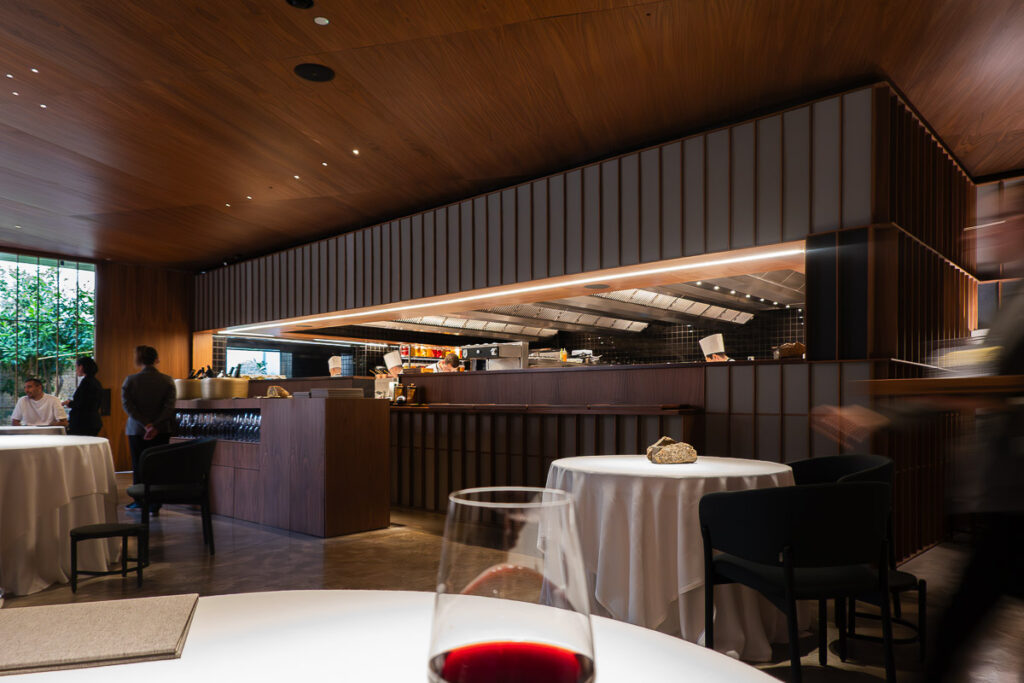 a glass of wine, circle tables and chefs cooking in the background which is the interior wooden dining area of Camarena restaurant in Valencia Spain