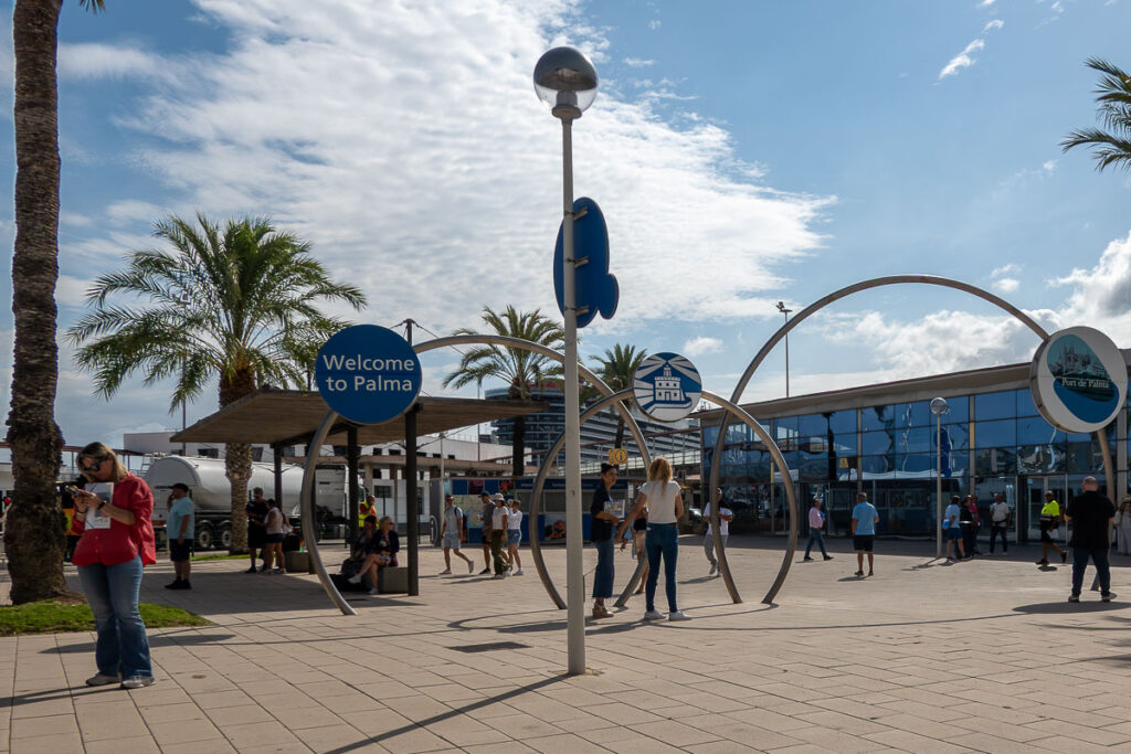 a plaza with steel arch sculpture and a building with a curtain glass in the background and people walking which is the Palma de Mallorca cruise terminal area